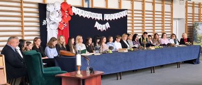 Sala, grupa osób siedzących za długim stołem, w tle dekoracja z biało czerwonych kwiatów z napisem „Narodowe Czytanie 2025”                                                                   