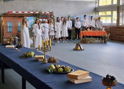 Sala, grupa dziewcząt w białych sukienkach, w tle chłopcy tworzący chór, na pierwszym planie długi stół z rekwizytami