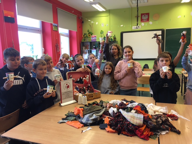 Sala lekcyjna. Przy stole z materiałami stoją dzieci i trzymają w ręku kolorowe lalki. Przed nimi walizka z lalkami.