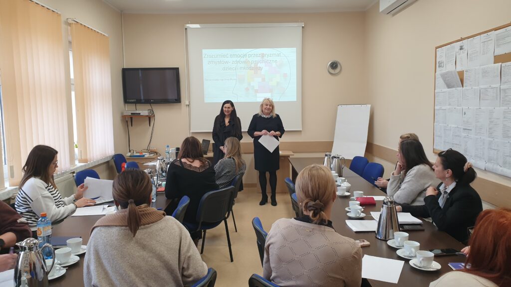 dwie kobiety stoją na tle wyświetlanej prezentacji grupa osób siedzi przy stolikach 