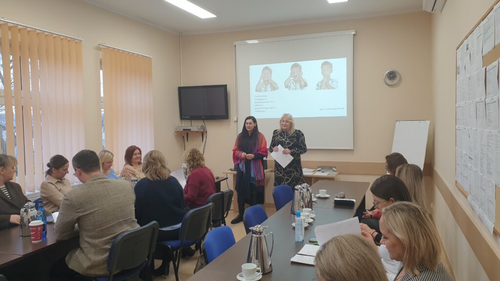 dwie kobiety stoją na tle wyświetlanej prezentacji grupa osób siedzi przy stolikach 