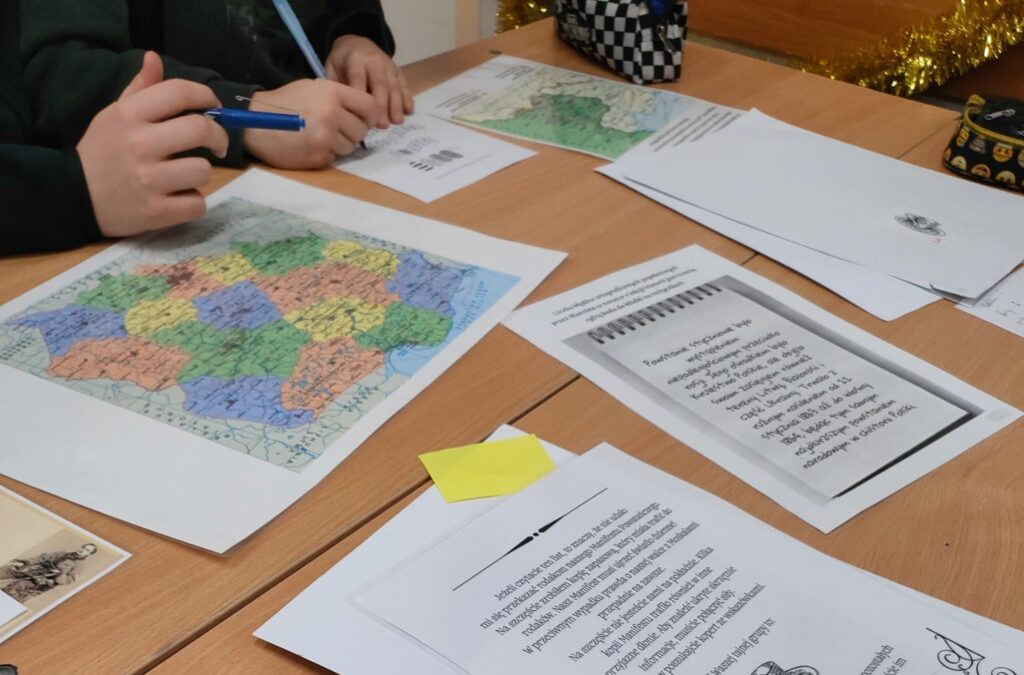 Na stole leżą kolorowe karty z mapami  i z zadaniami do escape roomu. W lewym, górnym rogu widać dłonie uczniów.