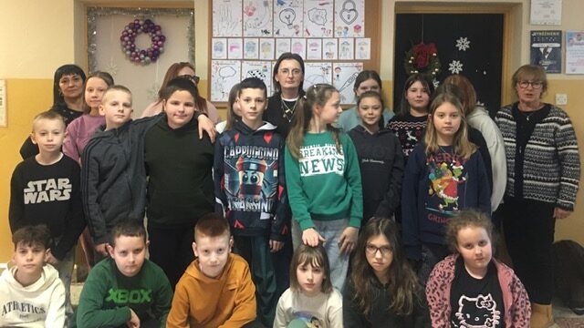 Korytarz szkolny, na tle kolorowej tablicy stoi duża grupa osób.