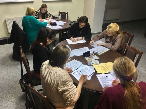 Sala na środku stół. Przy nim siedzą kobiety. Coś piszą. Na środku leżą białe kartki.