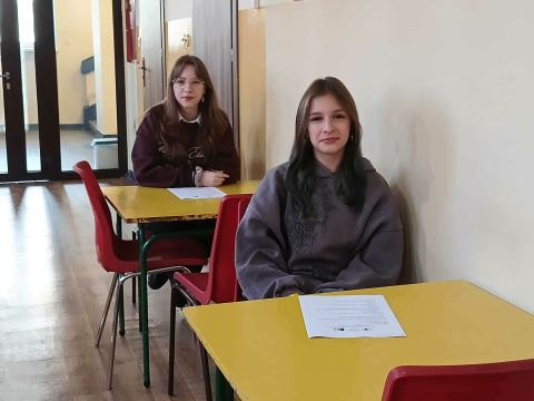 Hol szkoły, w przy stolikach siedzą dwie uczennice. Z prawej strony biała ściana.