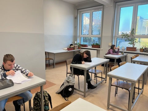 Sala lekcyjna, w ławkach siedzą uczniowie i piszą. 