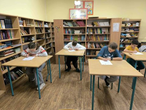 Biblioteka szkolna, przy stolikach siedzą uczniowie i piszą. 