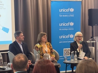 na fotelach siedzą trzy osoby przednimi soją okrągłe stoły za za jedną kobietą po prawej tronie stoi baner z napisem unicef