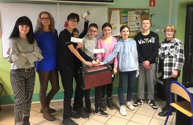 Sala lekcyjna, grupa 6 uczniów i trzy kobiety. Uczniowie trzymają walizkę i kartki.