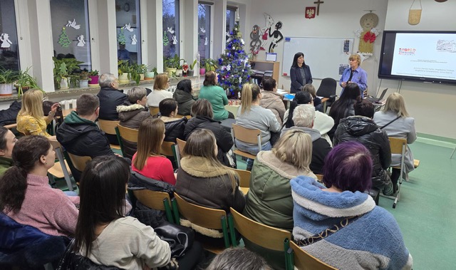 Sala szkolna, na krzesełkach siedzi bardzo dużo osób dorosłych. Przed nimi dwie kobiety stoją, prowadza spotkanie.
