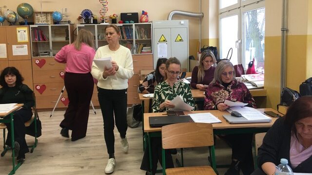 Sala lekcyjna. Przy stolikach siedzą uczestnicy. Mają na pulpicie kartki. Kobieta stoi na tle nauczycieli.