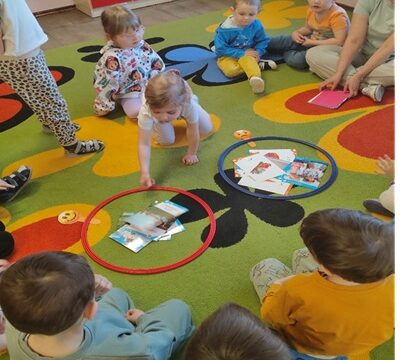 Dzieci z siedzą w kole na kolorowym dywanie. Przed sobą mają obręcze a w nich kartoniki z obrazkami. Jedna dziewczynka wskazuje obrazek.