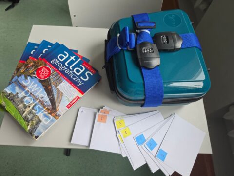 Na stoliku leży zamknięta na szyfr turkusowa walizka. Z boku leżą atlasy geograficzne i białe koperty z zadaniami