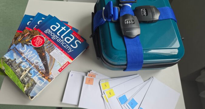 Na stoliku leży zamknięta na szyfr turkusowa walizka. Z boku leżą atlasy geograficzne i białe koperty z zadaniami