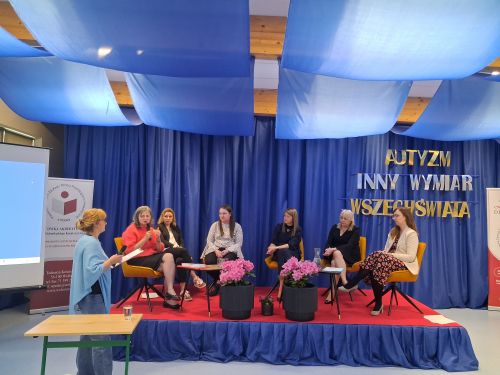 na środku sali na krzesłach siedzą kobiety przed nimi dwie czarne donice z kwiatami z boku stoi kobieta i prowadzi panel podczas konferencji 