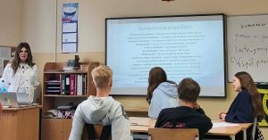 Sala lekcyjna, przy stolikach siedzą uczniowie. Przy biurku, naprzeciw nich stoi kobieta - prowadząca lekcję.